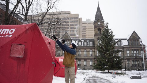 Des tentes devant l’Hôtel de Ville d’Halifax. Un homme vêtu d’un manteau utilise un balai pour enlever la neige du dessus de la tente. Paysage hivernal. 