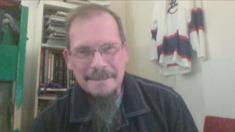 Un homme est assis à un bureau alors que derrière lui on distingue des livres et un chandail de hockey. Un homme est assis à un bureau alors que derrière lui on distingue des livres et un chandail de hockey.