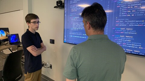 Un jeune homme debout les bras croisés regarde un homme plus vieux, de dos, debout devant un écran géant qui présente des lignes de code sur un fond bleu.