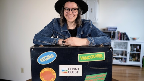 Isabelle Mercier souriante portant un chapeau noir et une veste en jean, appuyée sur une valise noire avec des autocollants de villes canadiennes.