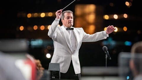 Jacques Lacombe, vêtu d'une veste blanche, tient une baguette de chef d'orchestre. 