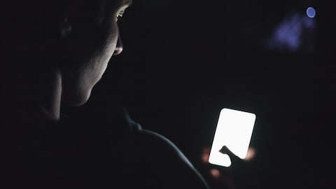 Un adolescent dans le noir, de dos, le visage penché sur un téléphone cellulaire allumé.