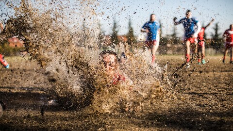 Un joueur de rugby s'éclabousse en tombant dans la boue, tandis qu'on voit d'autres joueurs à l'arrière.