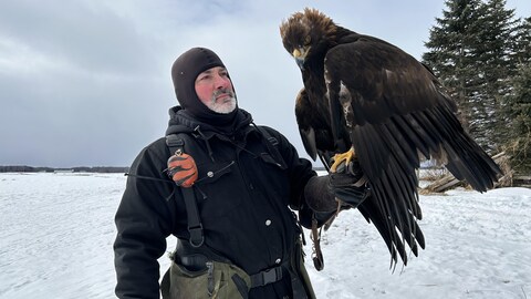 Un homme tient un grand oiseau de proie sur son bras, à l'extérieur, l'hiver. 