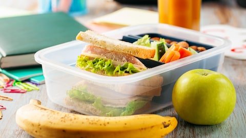 Un sandwich dans un plat de plastique, une banane et une pomme sur une table.