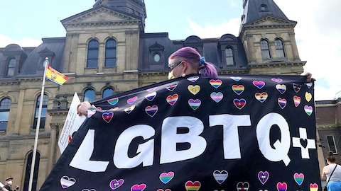 Une personne de dos, debout face à l'édifice de l'Assemblée législative du Nouveau-Brunswick, exhibant un grand drapeau noir décoré de l'acronyme LGBTQ+ en grosses lettres blanches et d'une dizaine de petits coeurs multicolores.