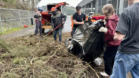 Des jeunes et un pompier versent des déchets verts devant une machine à déchiqueter et deux employés. 