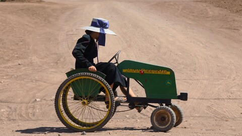 Une femme portant une robe traditionnelle mennonite et un chapeau gris au ruban bleu pédale sur un quadricycle vert de marque John Deere, sur une route de terre et de sable.