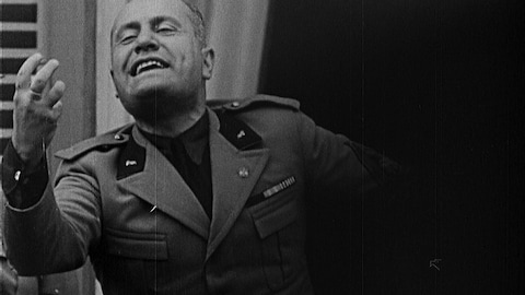 Benito Mussolini en survêtement militaire porte la main devant lui comme s'il allait attraper quelque chose.