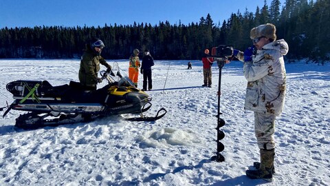 Un homme sur une motoneige regarde un autre homme percer des trous dans la glace à l'aide d'une perceuse à glace au lac Provencher de Baie-Comeau. 