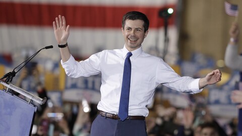 Pete Buttigieg salue la foule les bras ouverts sur scène près de sa tribune. 