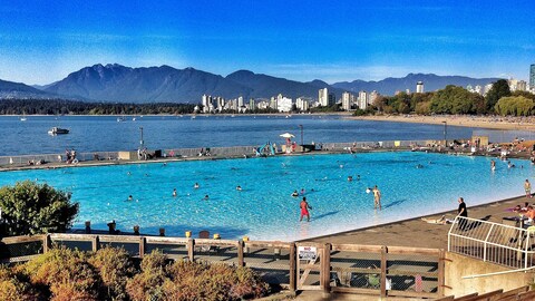 Des gens nagent dans une grande piscine extérieure avec vue sur le centre-ville de Vancouver, la plage, l'eau et les montagnes au loin.