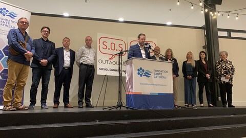 Les membres de la coalition SOS Outaouais sur une scène. Jean Pigeon qui est à la tête de la coalition, prend la parole au micro.