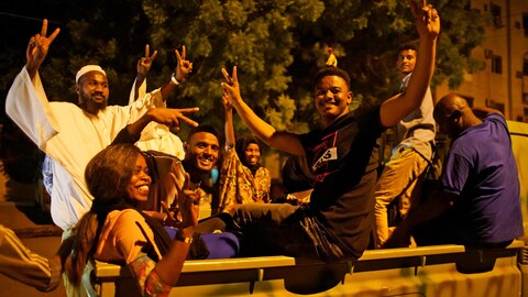 Plusieurs personnes font des signes de paix avec leurs doigts à l'arrière d'une camionnette dans la nuit à Khartoum.