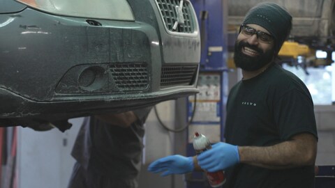 Un homme sourit dans un garage devant une voiture surélevée.
