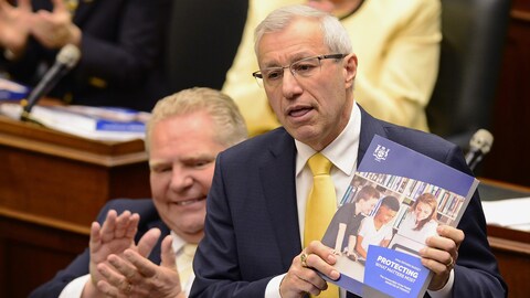 Le ministre en Chambre montrant un exemplaire du budget pendant que le premier ministre Doug Ford applaudit.