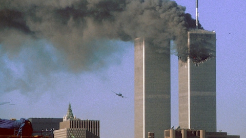 On voit les deux tours du World Trade Center, celle de droite est la proie d'un incendie et on y voit le trou béant résultant de l'impact du premier avion. À gauche, un appareil en vol se dirige sur la tour sud.  