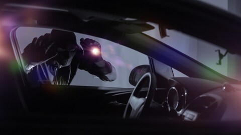 شخص مقنّع يحمل مصباحاً كهربائياً يحاول سرقة سيارة في الظلام. 