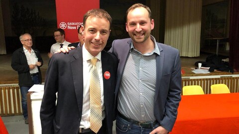 Yens Peterson et Ryan Meili sourient devant l'objectif devant une estrade où est installée une banière de Nouveau Parti démocratique. Yens Peterson et Ryan Meili sourient devant l'objectif devant une estrade où est installée une banière de Nouveau Parti démocratique.