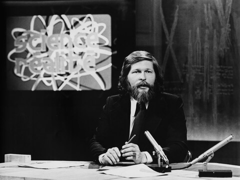 Dans un studio de télévision, l'animateur Joël Le Bigot est assis à une table
surmontée d'un micro. En arrière-plan, le titre de l'émission « Science Réalité » apparaît
dans un écran sur le mur. Dans un studio de télévision, l'animateur Joël Le Bigot est assis à une table
surmontée d'un micro. En arrière-plan, le titre de l'émission « Science Réalité » apparaît
dans un écran sur le mur.