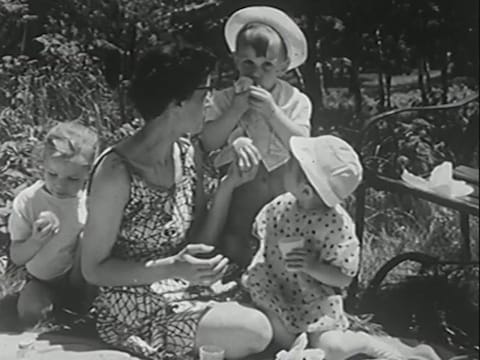 Mère accroupie sur une nappe de pique-nique, entourée de trois enfants qui mangent des fruits.