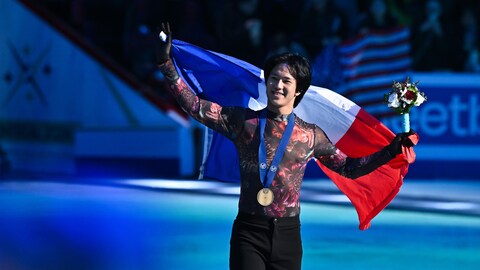 Un patineur sourit sur la glace avec une médaille de bronze au cou. Il tient un drapeau aux couleurs de la France.