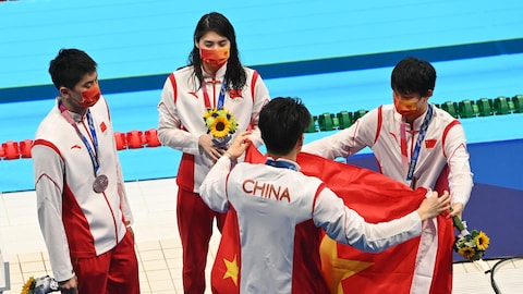 Quatre athlètes en survêtement sont sur le bord d'une piscine et déplient un drapeau de la Chine.