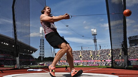 L'athlète, de profil, tient fermement à l'horizontal une boule accrochée à un fil de fer lors d'une rotation. 