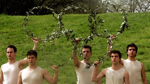 Cinq hommes forment les anneaux olympiques avec des couronnes de laurier qu'ils tiennent dans les airs.