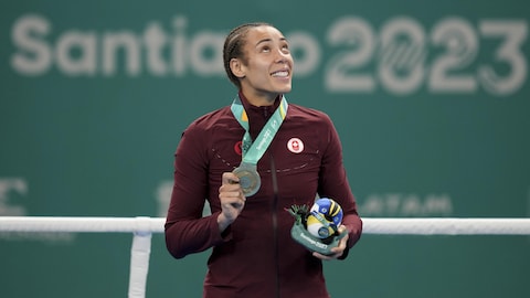 La boxeuse Tammara Thibeault tient avec sa main la médaille qu'elle a au cou en regardant vers le ciel sur le podium des Jeux panaméricains dans un ring devant le panneau Santiago 2023 