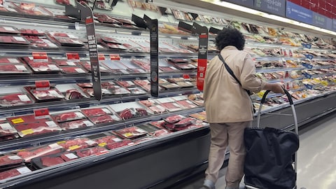 Une femme avec un panier se tient devant le comptoir des viandes dans un supermarché.
