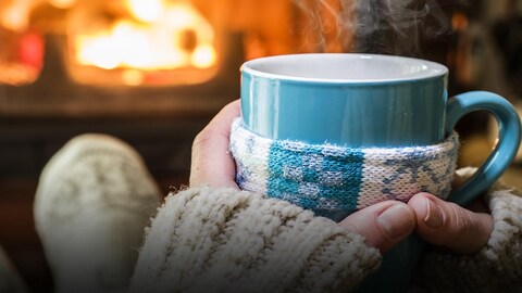 Une personne posant une tasse de café sur ses genoux, devant un feu de foyer.