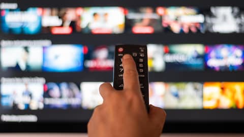On voit un écran en arrière plan avec une main au premier plan qui tient en main une télécommande.