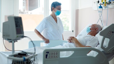 Une infirmière masquée parle à un patient alité dans une chambre d'hôpital. 