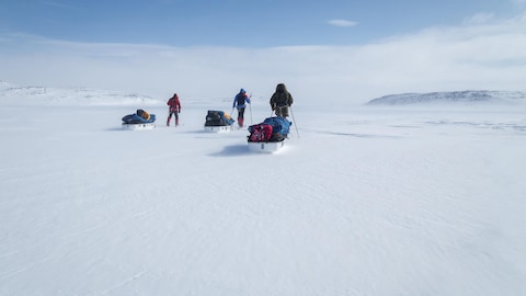 Trois personnes marchent dans une plaine enneigée avec des bagages dans des traîneaux. 