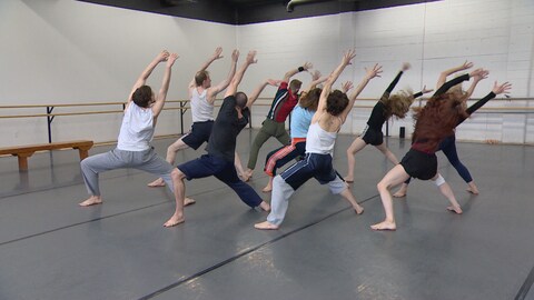 11 danseurs exécutant un mouvement les bras dans les air dans une salle de ballet au plancher gris et murs de briques blancs. Mai 2024.