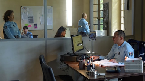 Plusieurs personnes dans un poste de police, dont une est assise à un bureau et regarde un ordinateur.