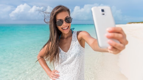 Une jeune femme portant lunettes soleil prend un autoportrait avec son téléphone, devant une plage et la mer bleue des Caraibes.