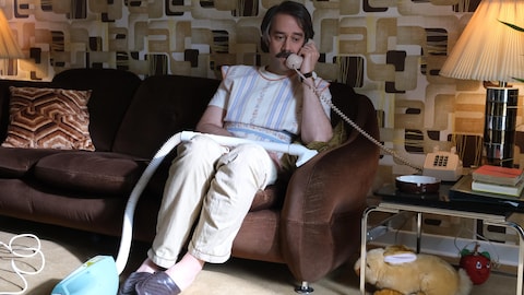 Gaétan Delisle (François Létourneau) avec son tablier et sa balayeuse, parle au téléphone dans le salon.