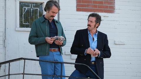 Gaétan (François Létourneau) et Serge (Patrice Robitaille) sont à l'extérieur, l'un compte son argent et l'autre fume une cigarette.
