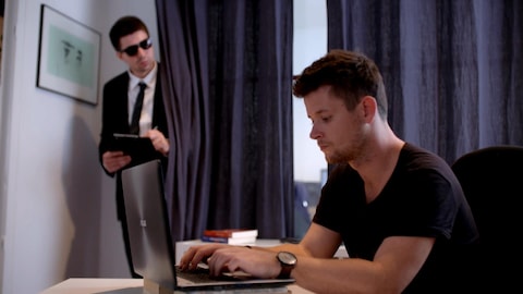 Un homme derrière son ordinateur se fait espionner par un courtier en données caché derrière un rideau.