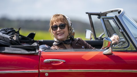 L'actrice Lesley Manville dans le rôle de Susan. Elle est dans une voiture décapotable et porte des verres fumés et un foulard.