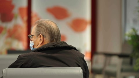 Un homme agé porte un masque, assis sur une chaise dans la salle d'attente d'une clinique.