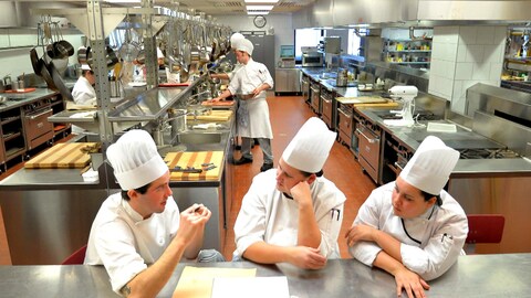Trois chefs attablés à un comptoir en inox dans une grande cuisine industrielle. 