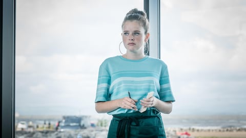 Une jeune femme devant une baie vitrée tient un carnet avec un air désespéré et triste.