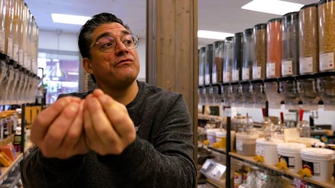 Radio-Canada Un épicier montre quelque chose avec ses mains entouré d'aliments en vrac dans un marché.