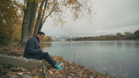 Un homme assis dehors en train d'écrire. Il est au bord de l'eau.