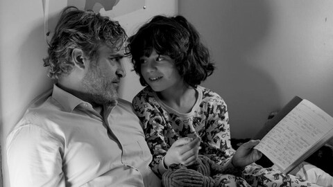 En noir et blanc, un homme (Joaquin Phoenix) installé dans le lit d'un enfant regarde ce dernier.