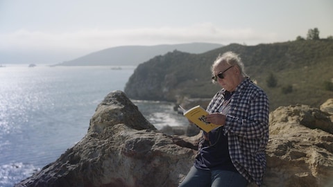 Lucien Francoeur lit un livre dehors, appuyé sur un rocher.