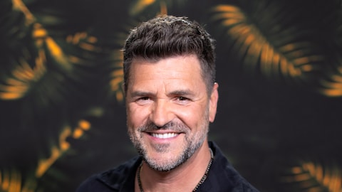 Le chanteur sourit à la caméra, devant un décor tropical.